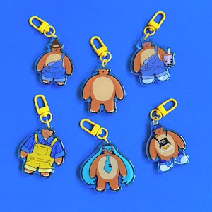 May include: A collection of colorful bear-shaped keychains with yellow clasps. Each keychain features a unique bear design, including bears wearing overalls, hats, and holding a drink. The keychains are set against a solid blue background.