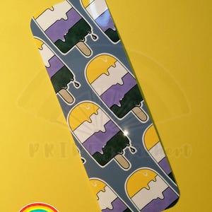 Könnte beinhalten: Ein Lesezeichen mit einem Muster aus gelben, weißen, lila und grünen Eis am Stiel auf blauem Hintergrund. Das Lesezeichen ist aus glänzendem Papier und hat ein Regenbogenlogo mit dem Text "PRIDE gefeiert" unten.