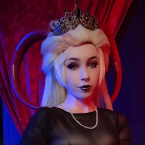 Può includere: Una donna indossa un top nero trasparente con un abito di paillettes viola e una corona. Tiene un serpente tra le mani. Lo sfondo è rosso e blu.