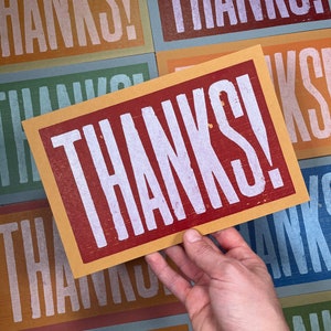 A hand holds an oversized orange card with maroon ink and the word Thanks! in white ink with a background of all the different colored card options that also read Thanks!.