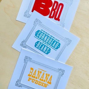 May include: Three white cards with decorative black frames and colorful text. The top card says "BBQ" in red letters. The middle card says "Feed me on cornbread and beans" in teal letters. The bottom card says "Banana Puddin'" in yellow letters.
