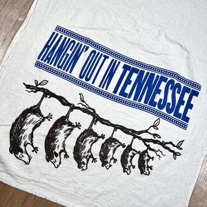 May include: White kitchen towel with a blue rectangular text box that reads "HANGIN' OUT IN TENNESSEE". Below the text is a black illustration of several opossums hanging from a tree branch. The towel is made of a textured fabric.