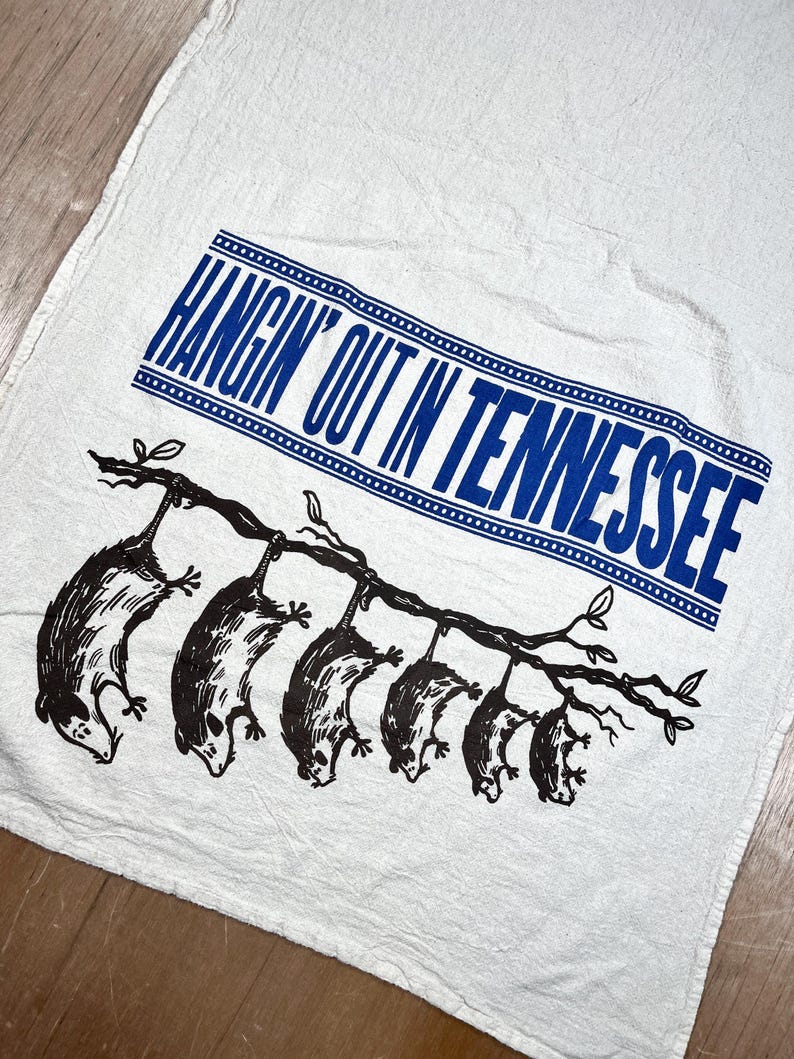May include: White kitchen towel with a blue rectangular text box that reads "HANGIN' OUT IN TENNESSEE". Below the text is a black illustration of several opossums hanging from a tree branch. The towel is made of a textured fabric.
