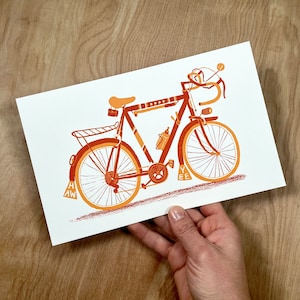 A hand printed letterpress illustration of a vintage style 10-speed road racer bicycle, featuring drop handlebars and mudflaps, in orange and red inks on bright white cardstock, photographed on a wood background.