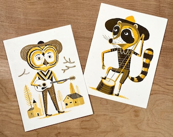 Puede incluir: Dos impresiones ilustradas con personajes animales. La primera impresión muestra un búho con un sombrero de vaquero tocando la guitarra. La segunda impresión muestra un mapache con un sombrero de vaquero sosteniendo un palo. Ambas impresiones tienen una gama de colores amarillo y marrón.
