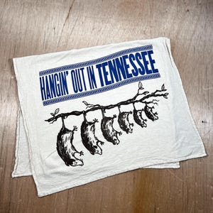 May include: Off-white, rectangular kitchen towel with the text "HANGIN' OUT IN TENNESSEE" in blue. Below the text, a branch with six black opossums hanging upside down is printed on the towel.