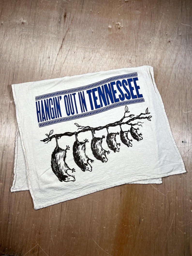 May include: Off-white, rectangular kitchen towel with the text "HANGIN' OUT IN TENNESSEE" in blue. Below the text, a branch with six black opossums hanging upside down is printed on the towel.