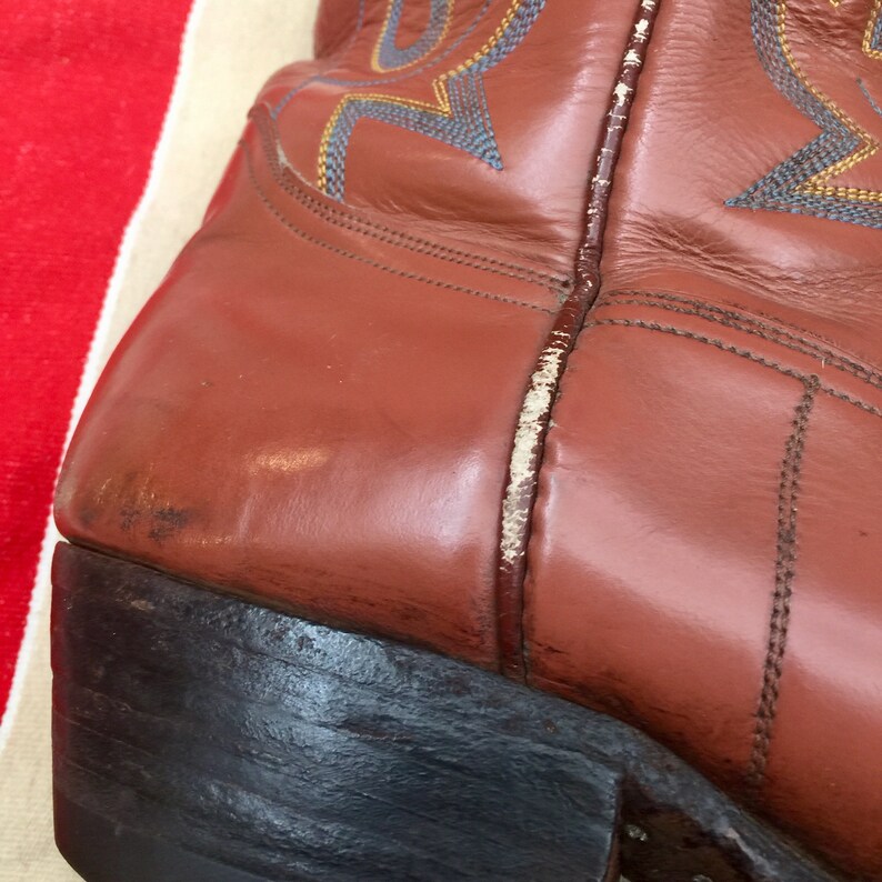 May include: Close-up of the side of a brown leather cowboy boot with a white stitching detail. The boot has a black leather sole.
