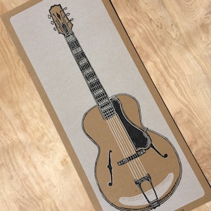 A black and white illustrated hand carved relief print of an archtop guitar on kraft colored paper lays on a wood grain background.