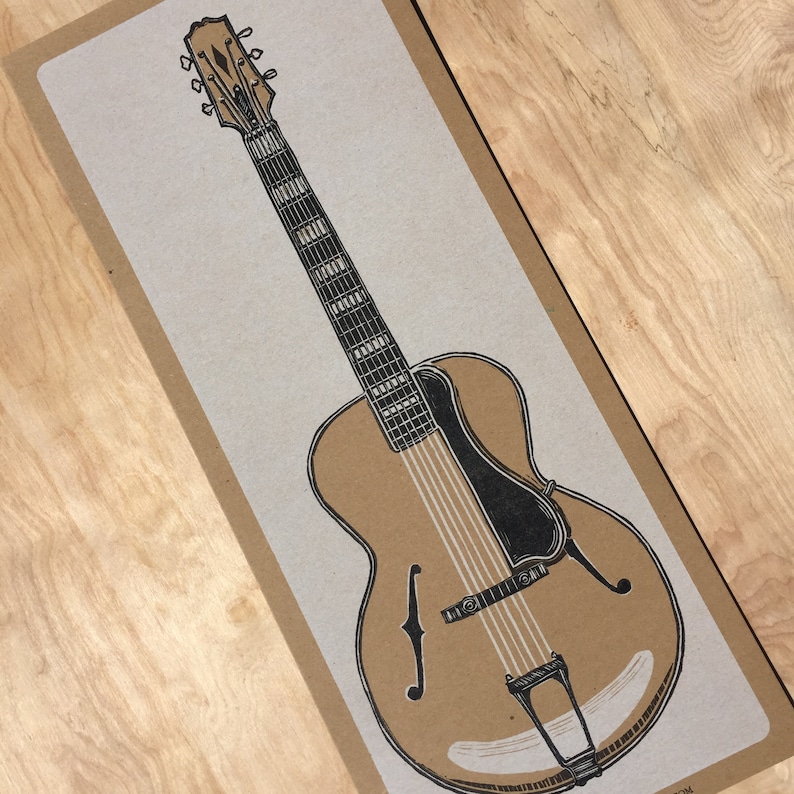 A black and white illustrated hand carved relief print of an archtop guitar on kraft colored paper lays on a wood grain background.