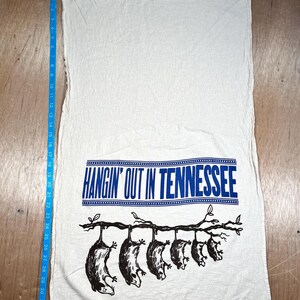 May include: White kitchen towel with the text "HANGIN' OUT IN TENNESSEE" in blue. Below the text is a black illustration of opossums hanging from a tree branch. The towel measures approximately 34 inches long.