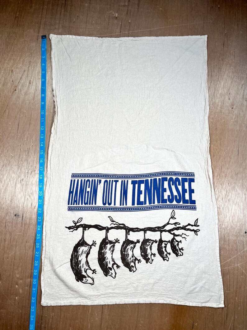 May include: White kitchen towel with the text "HANGIN' OUT IN TENNESSEE" in blue. Below the text is a black illustration of opossums hanging from a tree branch. The towel measures approximately 34 inches long.