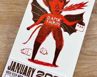 May include: A January 2027 calendar page featuring a red and brown illustration of a horned devil character with wings, breathing fire, and holding a jug labeled XXX. The text DAMN THIRSTY is printed on the chest, with additional jugs on the ground.