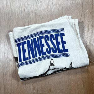 May include: Folded white kitchen towel with the word "TENNESSEE" in large blue letters. The towel has a dotted border and a black branch design. The towel is folded on a wooden surface.