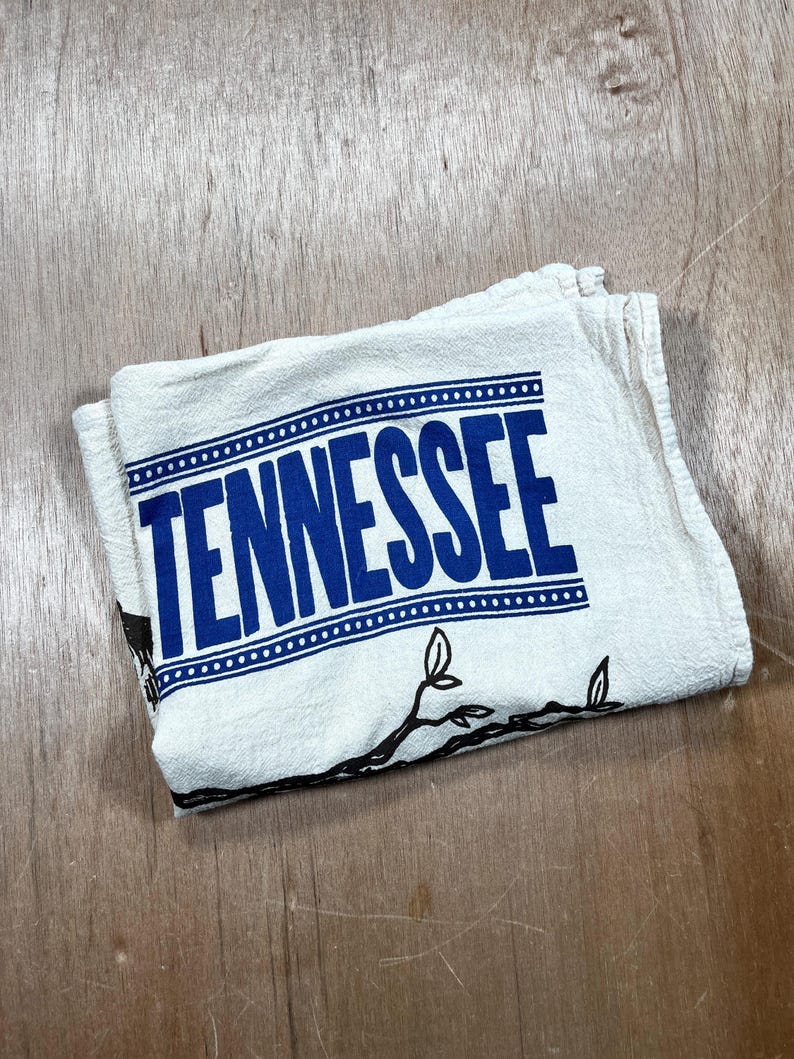 May include: Folded white kitchen towel with the word "TENNESSEE" in large blue letters. The towel has a dotted border and a black branch design. The towel is folded on a wooden surface.
