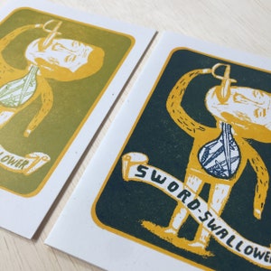 May include: Two illustrated cards with a yellow figure with swords sticking out of its body. The card on the left has a green background and the text "FLOWER" on a scroll. The card on the right has a dark green background and the text "SWORD-SWALLOWER" on a scroll.