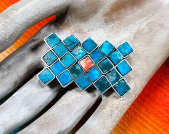 May include: A silver ring featuring a geometric design of turquoise and coral-colored stones. The ring's stones are square-cut and set in a grid pattern, creating a striking visual effect. The ring is displayed on a hand.