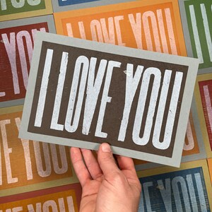 A hand holds an oversized light blue card with brown ink and the words I Love You in white ink with a background of all the different colored card options that also read I Love You.