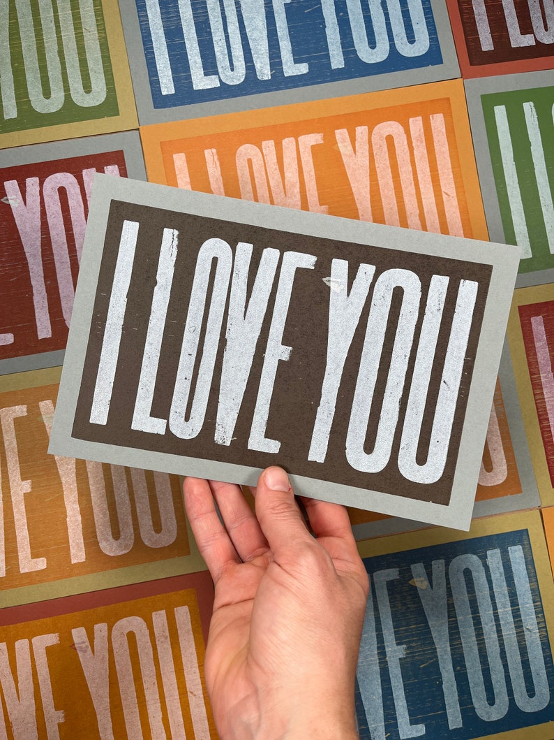 A hand holds an oversized light blue card with brown ink and the words I Love You in white ink with a background of all the different colored card options that also read I Love You.