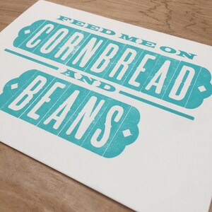 May include: A teal blue and white print with the text "Feed me on cornbread and beans".