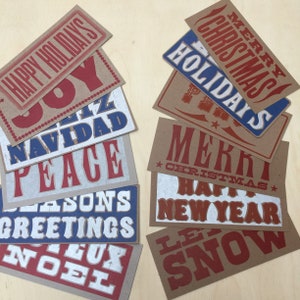 May include: A collection of eight brown paper cards with red and blue lettering. The cards feature various holiday greetings, including "Happy Holidays", "Joy", "Feliz Navidad", "Peace", "Seasons Greetings", "Noel", "Merry Christmas", "Holidays", "Merri Christmas", "Happy New Year", and "Let It Snow".