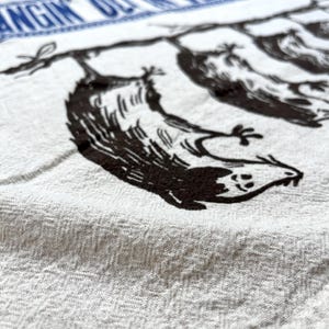 May include: White kitchen towel with a blue border featuring the text "Hangin' Out in the Kitchen". The towel has a black illustration of birds hanging from a branch. The towel is made of a textured fabric.