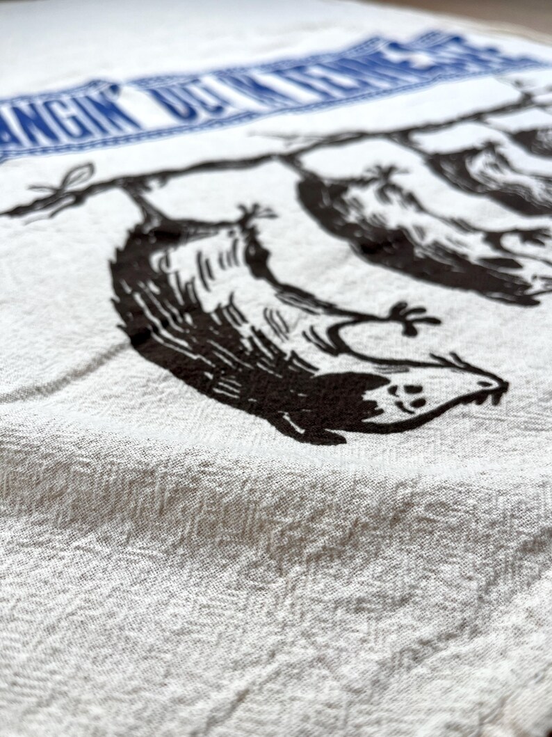 May include: White kitchen towel with a blue border featuring the text "Hangin' Out in the Kitchen". The towel has a black illustration of birds hanging from a branch. The towel is made of a textured fabric.