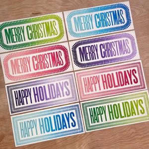 May include: A collection of eight rectangular labels featuring decorative borders. Four labels display the text Merry Christmas in green, blue, red, and purple. Four labels display the text Happy Holidays in purple, red, blue, and green ink colors.