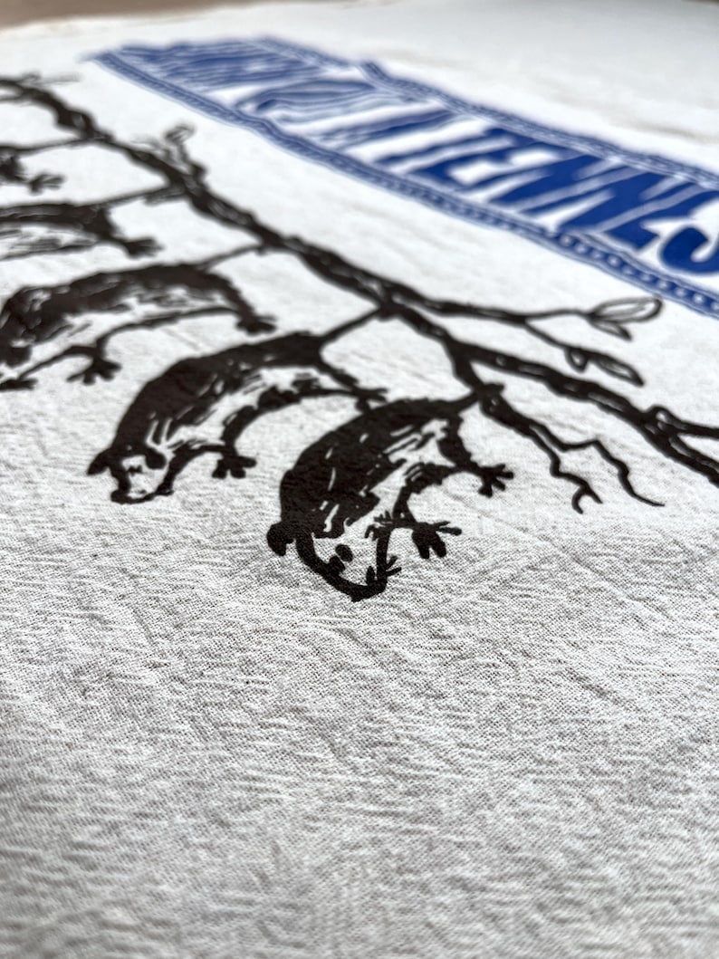 May include: A close-up of a white textile with a black illustration of possums on tree branches. The textile has a blue rectangular border with white text. The possums are in a stylized, illustrative style.