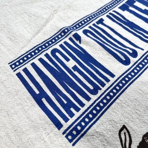 May include: White linen towel with a blue border and the words "HANGIN' OUT" printed in blue. The towel also features a black floral design in the lower right corner.