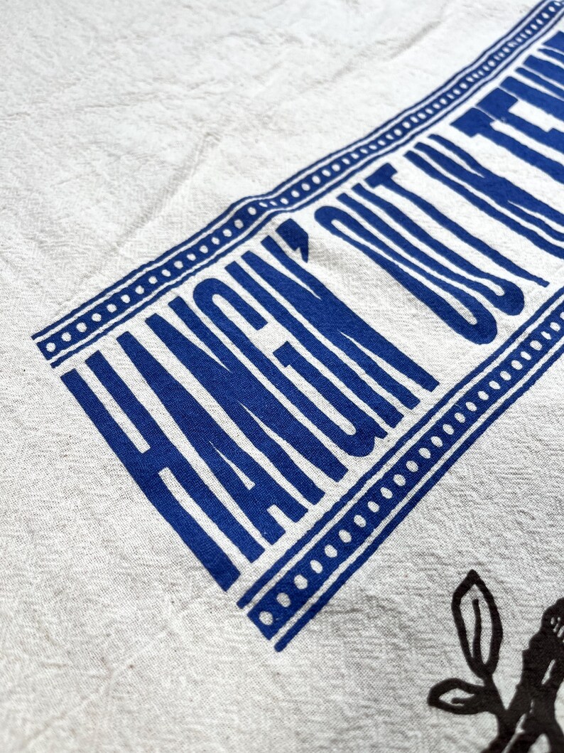 May include: White linen towel with a blue border and the words "HANGIN' OUT" printed in blue. The towel also features a black floral design in the lower right corner.