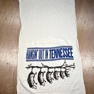 May include: Off-white kitchen towel with the text "HANGIN' OUT IN TENNESSEE" in blue. Below the text, a branch with six opossums hanging from it is printed in black. The towel is made of a soft, textured fabric.
