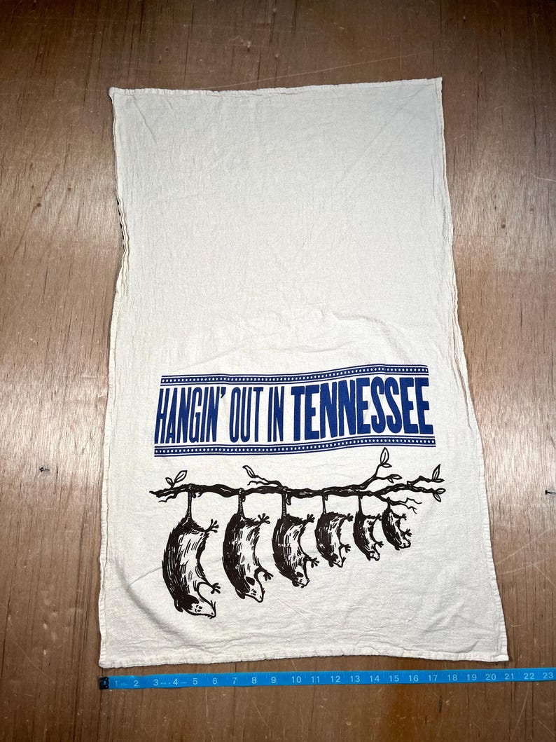 May include: Off-white kitchen towel with the text "HANGIN' OUT IN TENNESSEE" in blue. Below the text, a branch with six opossums hanging from it is printed in black. The towel is made of a soft, textured fabric.