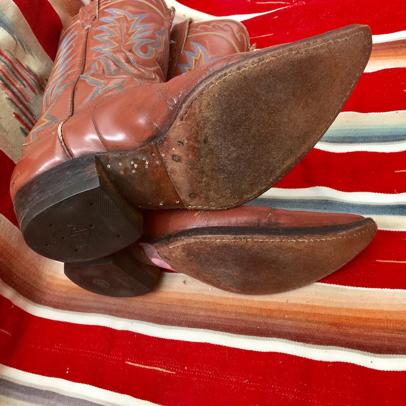May include: A pair of brown leather cowboy boots with intricate blue and orange stitching on the shaft. The boots are worn and have a distressed look. The soles are brown leather and have a slight curve.