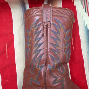 May include: Brown leather cowboy boots with blue and yellow stitching. The boots have a pointed toe and a tall shaft.