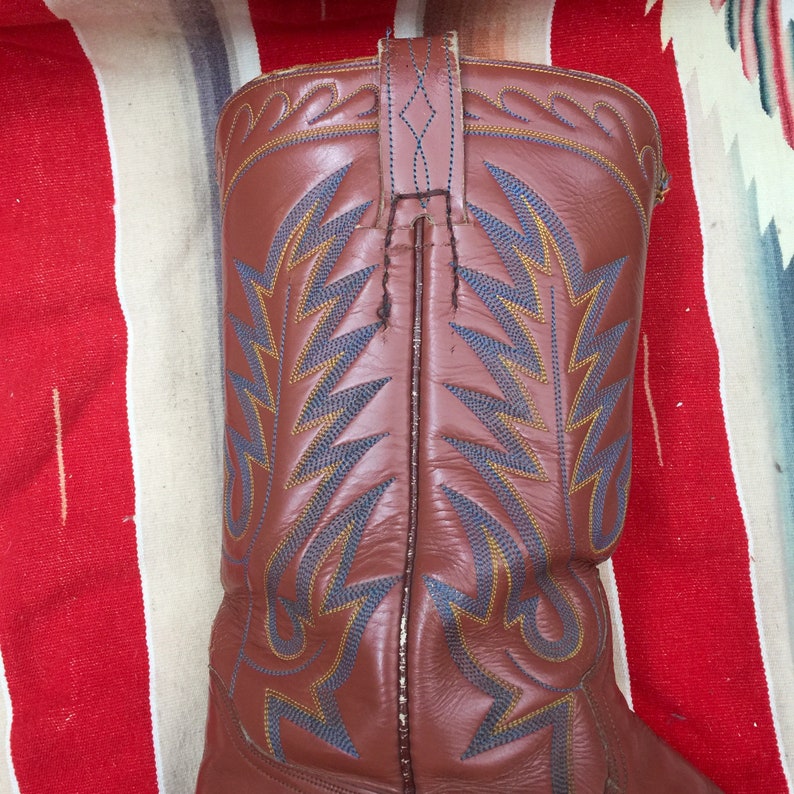 May include: Brown leather cowboy boots with blue and yellow stitching. The boots have a pointed toe and a tall shaft.