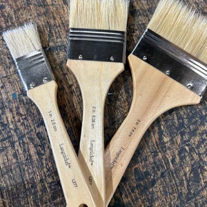 May include: Three paint brushes with natural bristles and wooden handles. The brushes are labeled with their sizes: 1 inch, 2 inches, and 3 inches. The brand name "Langnickel" is printed on each handle.