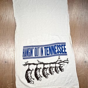 May include: White kitchen towel with a blue banner that reads "HANGIN' OUT IN TENNESSEE." Below the banner, a black illustration depicts several opossums hanging from a tree branch. The towel is made of a soft, textured fabric.
