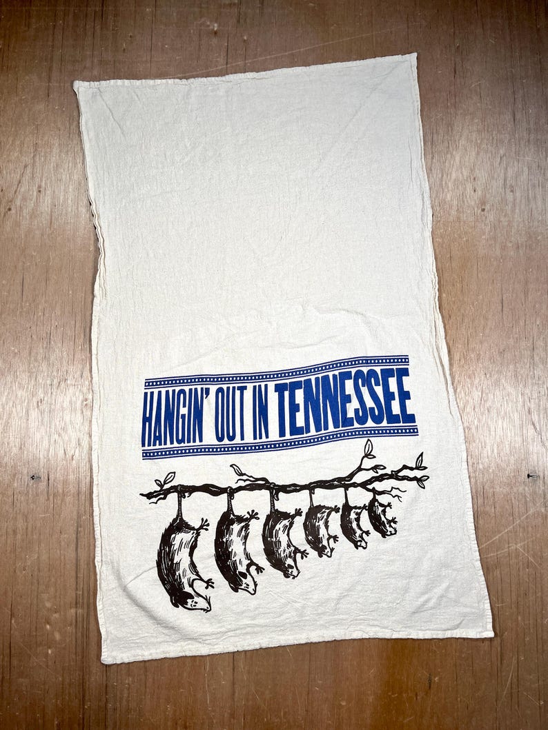 May include: White kitchen towel with a blue banner that reads "HANGIN' OUT IN TENNESSEE." Below the banner, a black illustration depicts several opossums hanging from a tree branch. The towel is made of a soft, textured fabric.