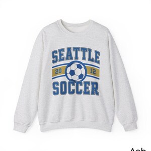 Pode incluir: Camisola cinza-claro com a inscri&ccedil;&atilde;o "SEATTLE SOCCER" em azul, um gr&aacute;fico de bola de futebol e os n&uacute;meros "20" e "12". A camisola tem gola redonda.