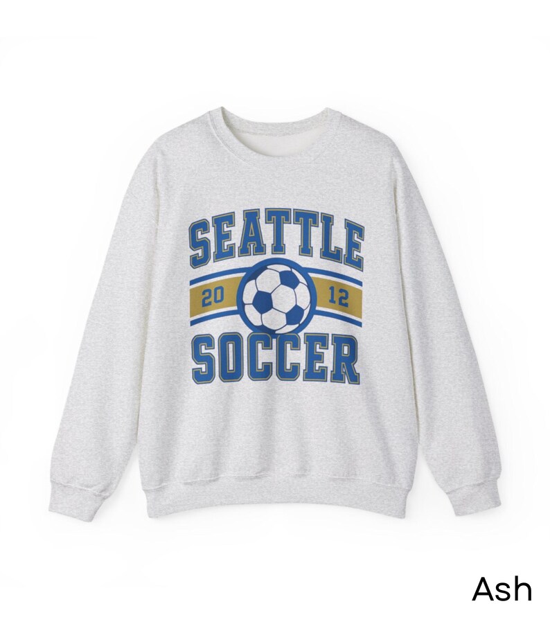 Pode incluir: Camisola cinza-claro com a inscri&ccedil;&atilde;o "SEATTLE SOCCER" em azul, um gr&aacute;fico de bola de futebol e os n&uacute;meros "20" e "12". A camisola tem gola redonda.