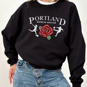 May include: Black sweatshirt with a white graphic print that says "PORTLAND WOMENS SOCCER" with a red rose and two football players.