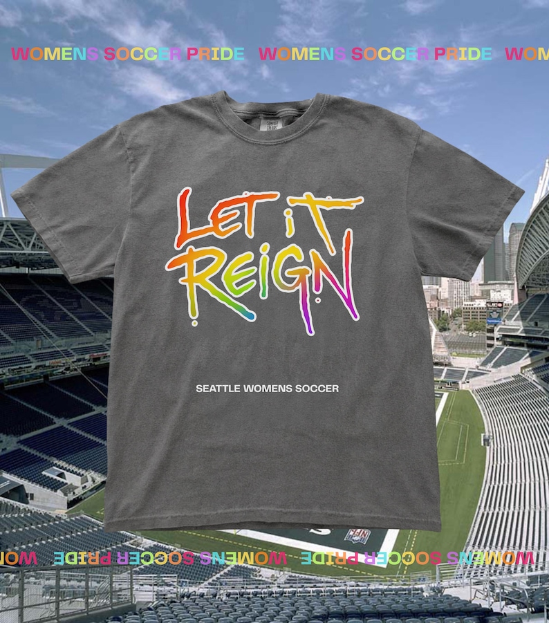 May include: A gray t-shirt with the words "LET IT REIGN" in a rainbow gradient design. Below the text is "SEATTLE WOMENS SOCCER" in white. The shirt is displayed against a backdrop of a stadium.