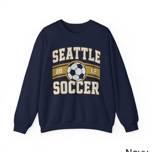 Pode incluir: Camisola azul marinho com o texto "SEATTLE SOCCER" e um gr&aacute;fico de bola de futebol. O design inclui os n&uacute;meros "20" e "12". A palavra "Navy" est&aacute; impressa no canto inferior direito.