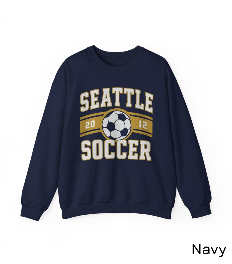 Pode incluir: Camisola azul marinho com o texto "SEATTLE SOCCER" e um gr&aacute;fico de bola de futebol. O design inclui os n&uacute;meros "20" e "12". A palavra "Navy" est&aacute; impressa no canto inferior direito.