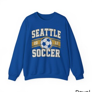 Pode incluir: Camisola azul royal com o texto "SEATTLE SOCCER" em letras brancas. Um gr&aacute;fico de bola de futebol est&aacute; centrado acima da palavra "SOCCER". Os n&uacute;meros "20" e "12" est&atilde;o em ambos os lados do gr&aacute;fico da bola de futebol.