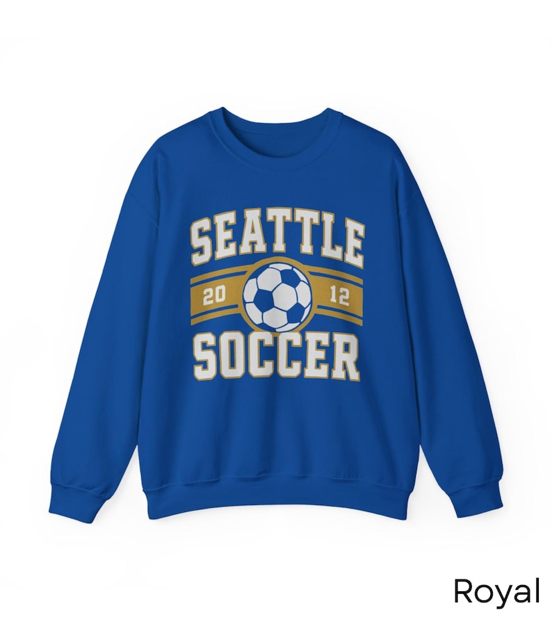 Pode incluir: Camisola azul royal com o texto "SEATTLE SOCCER" em letras brancas. Um gr&aacute;fico de bola de futebol est&aacute; centrado acima da palavra "SOCCER". Os n&uacute;meros "20" e "12" est&atilde;o em ambos os lados do gr&aacute;fico da bola de futebol.
