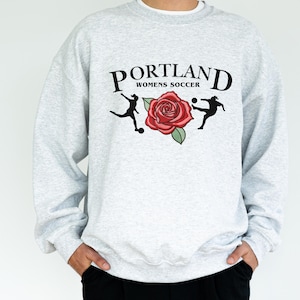 May include: A grey sweatshirt with a graphic print of a rose and two football players. The text "PORTLAND WOMENS SOCCER" is printed above the graphic.