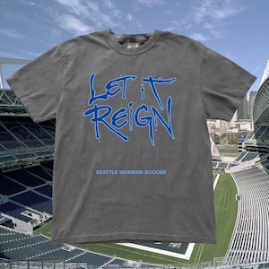 May include: A gray t-shirt with the words "LET IT REIGN" in blue, above the text "SEATTLE WOMENS SOCCER." The shirt is displayed against a backdrop of a soccer stadium and a blue sky.
