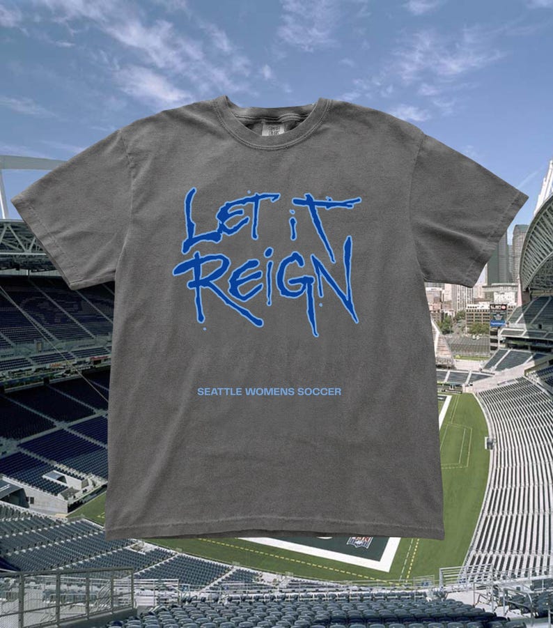 May include: A gray t-shirt with the words "LET IT REIGN" in blue, above the text "SEATTLE WOMENS SOCCER." The shirt is displayed against a backdrop of a soccer stadium and a blue sky.